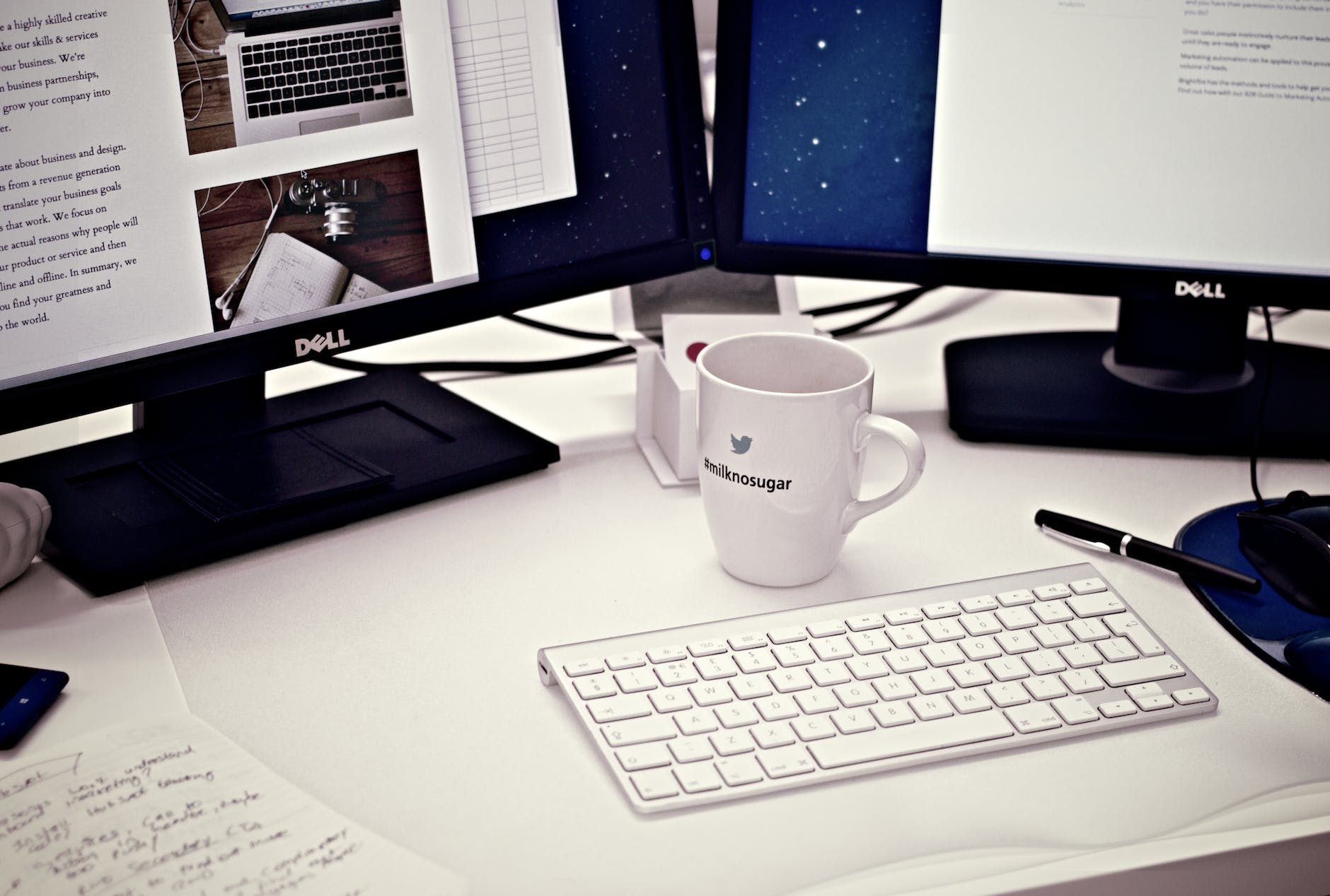 Partial view of two Dell monitors with text and images on them  on a white desk with a coffee mug in the middle below them, a black pen and mousepad with a partial picture of a mouse, and what looks like an Apple keyboard below the coffee mug and papers to the left with scribble notes