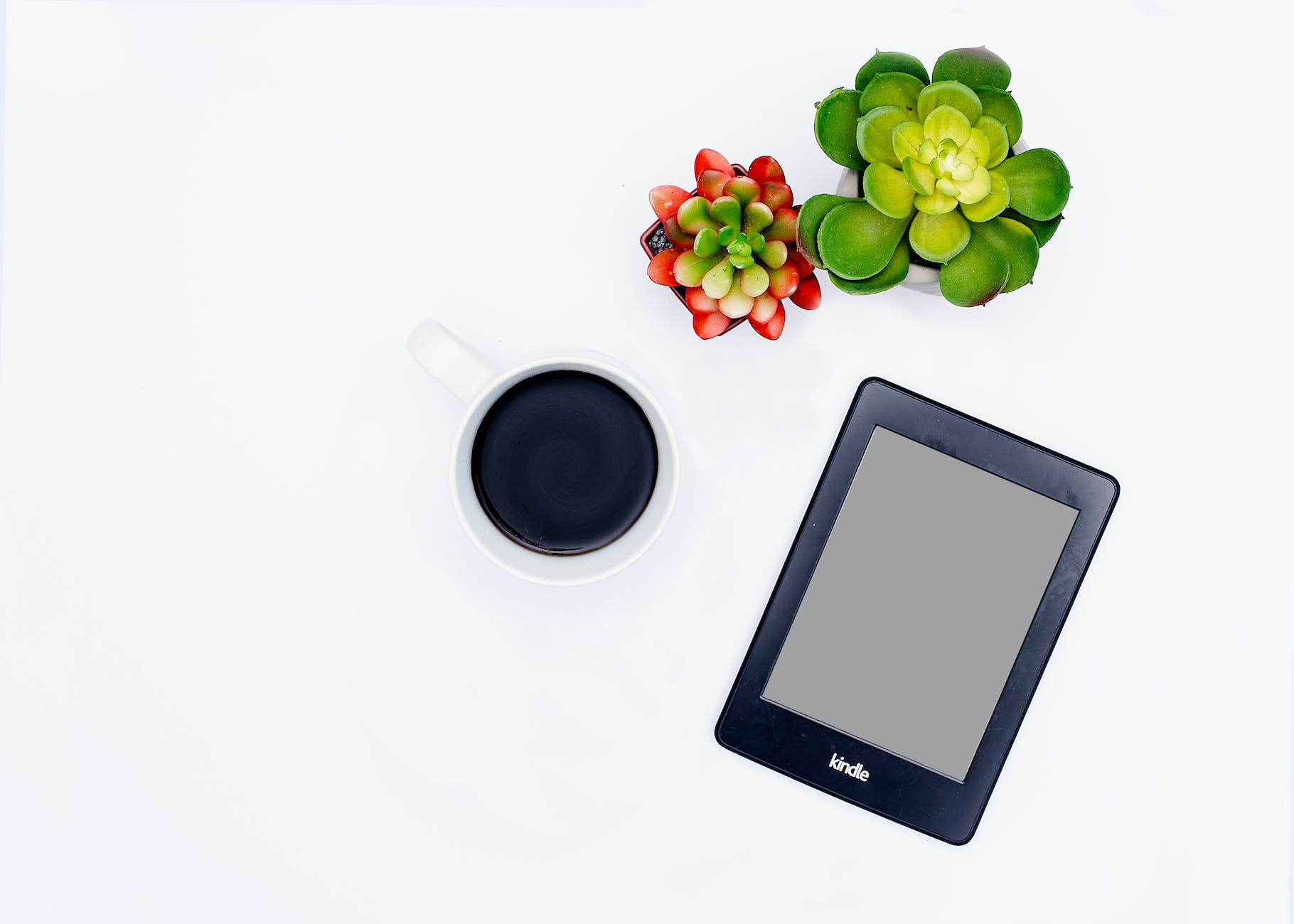 Top-down look at a white coffee mug with coffee in the center, a Kindle tilted 45 degrees to the bottom right with a red and green small-leaf succulent and a larger large-leaf succulent immediately to the right and above the Kindle on a white background.